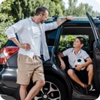 Father watches as son climbs out of back seat of a vehicle holding a soccer ball.